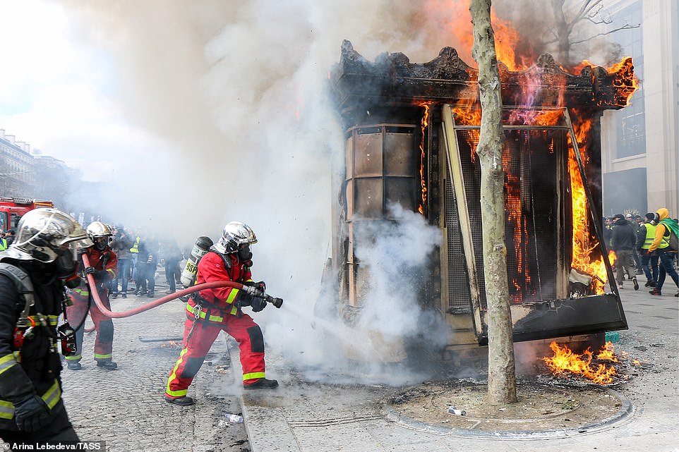 Firefighters battle a fire at a street kiosk during a Yellow Vests movement protest as demonstrations continue around them