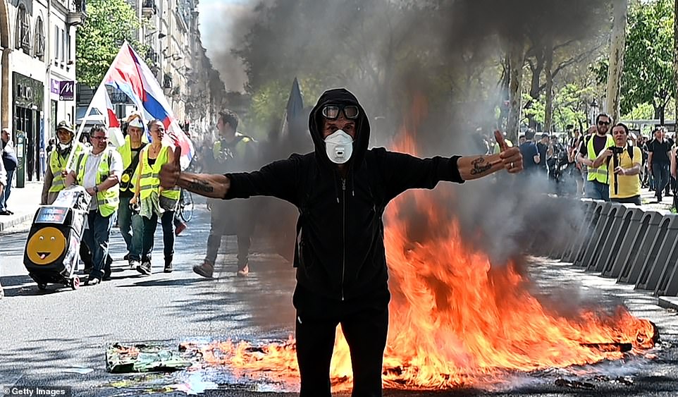 A protester from the yellow vests stands in front of a burning bin today and gives the cameras two thumbs up