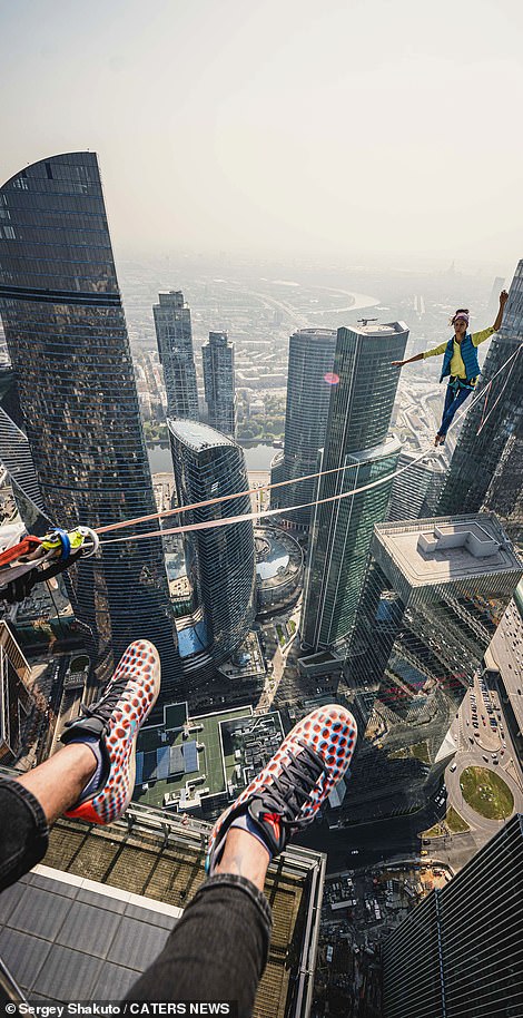 One of the athletes dangles his feet over the edge of the skyscraper as another walks along the slack line